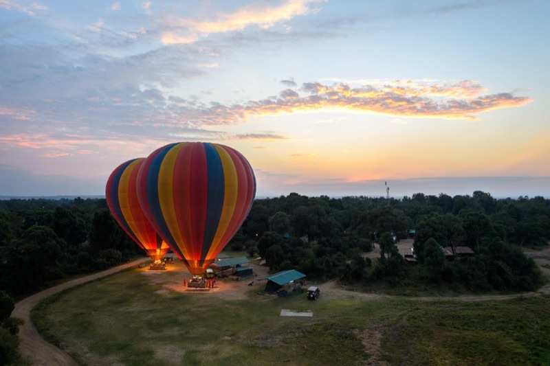 Two hot-air balloons standing inflated and lit at dawn on a clearing beside a camp, seen from above