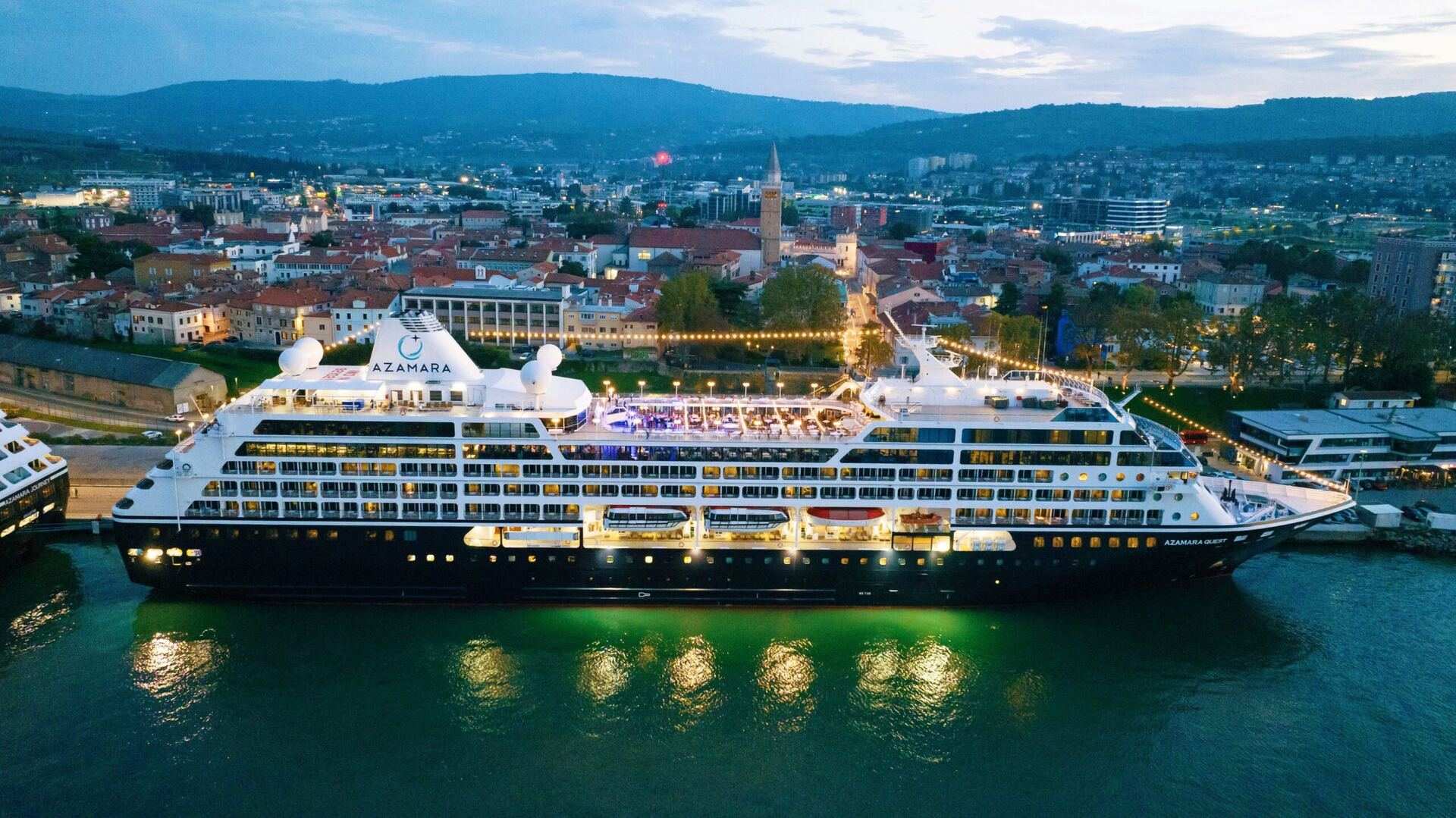Azamara Quest lit up at dusk, moored along the quay of an old harbour town