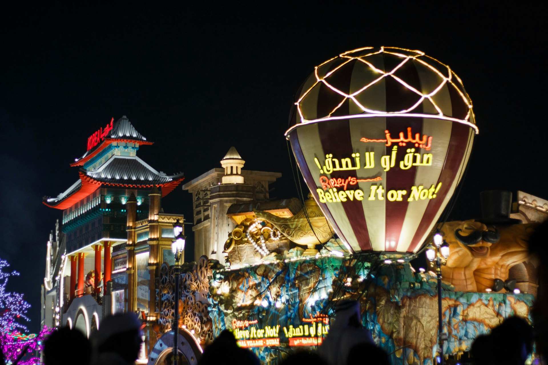 Lit pavilions and a glowing balloon-shaped sign at Global Village in Dubai at night, with a crowd in silhouette in front