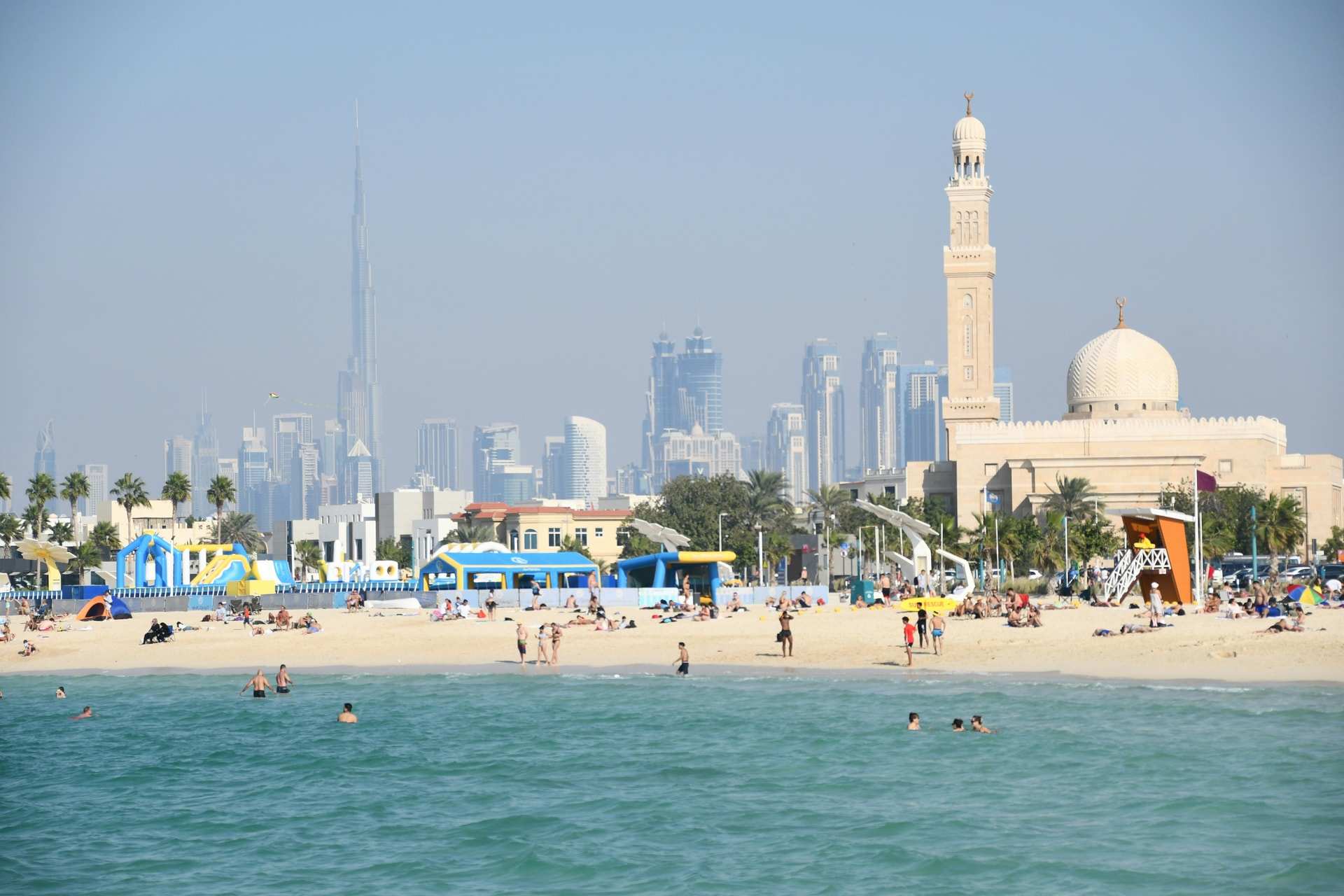 Swimmers in calm green water off a busy public beach in Dubai, with a white mosque and minaret and the Downtown towers behind in the haze
