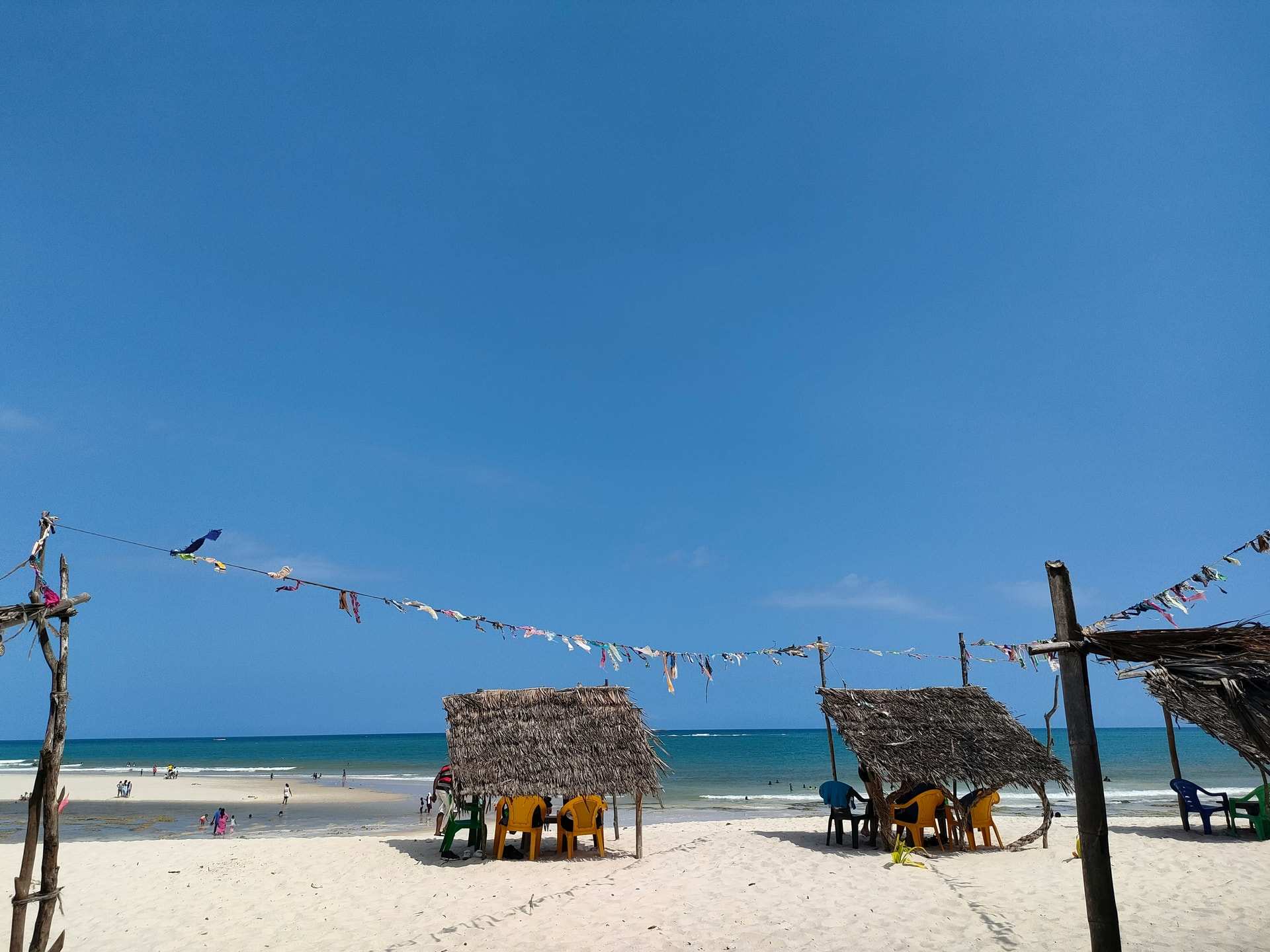 Thatched beach shelters with blue and yellow chairs on white sand at Diani, with bunting strung between poles and a calm sea behind