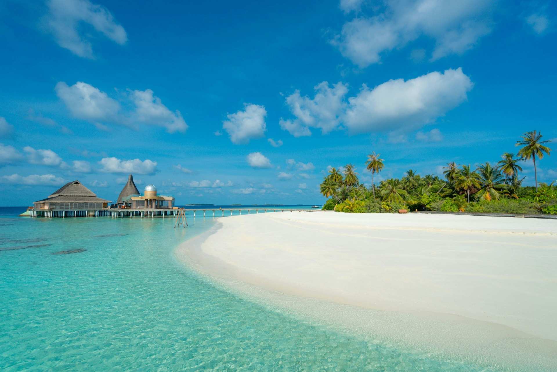A white sand spit curving out into a shallow turquoise lagoon beside a resort island in Baa Atoll, with thatched buildings on the water