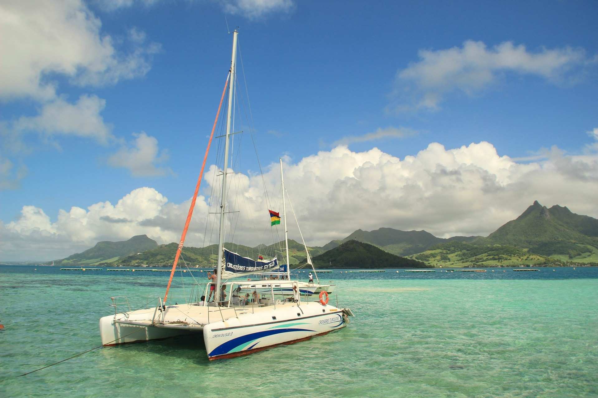 A catamaran at anchor in a pale green lagoon off Mauritius, with a Mauritian flag at the stern and green hills behind