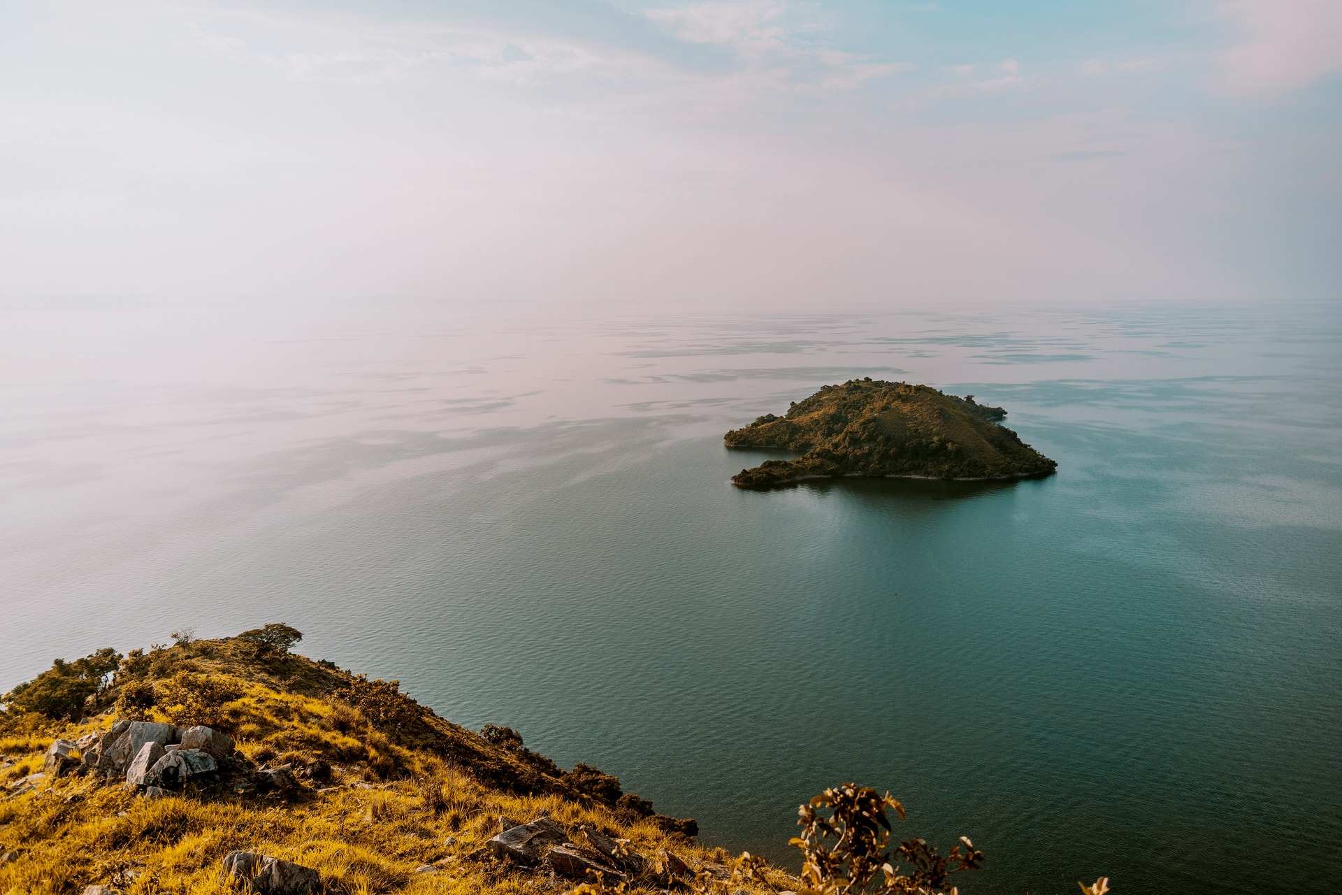 A small wooded island in the calm, hazy water of Lake Kivu, seen from a grassy rocky point
