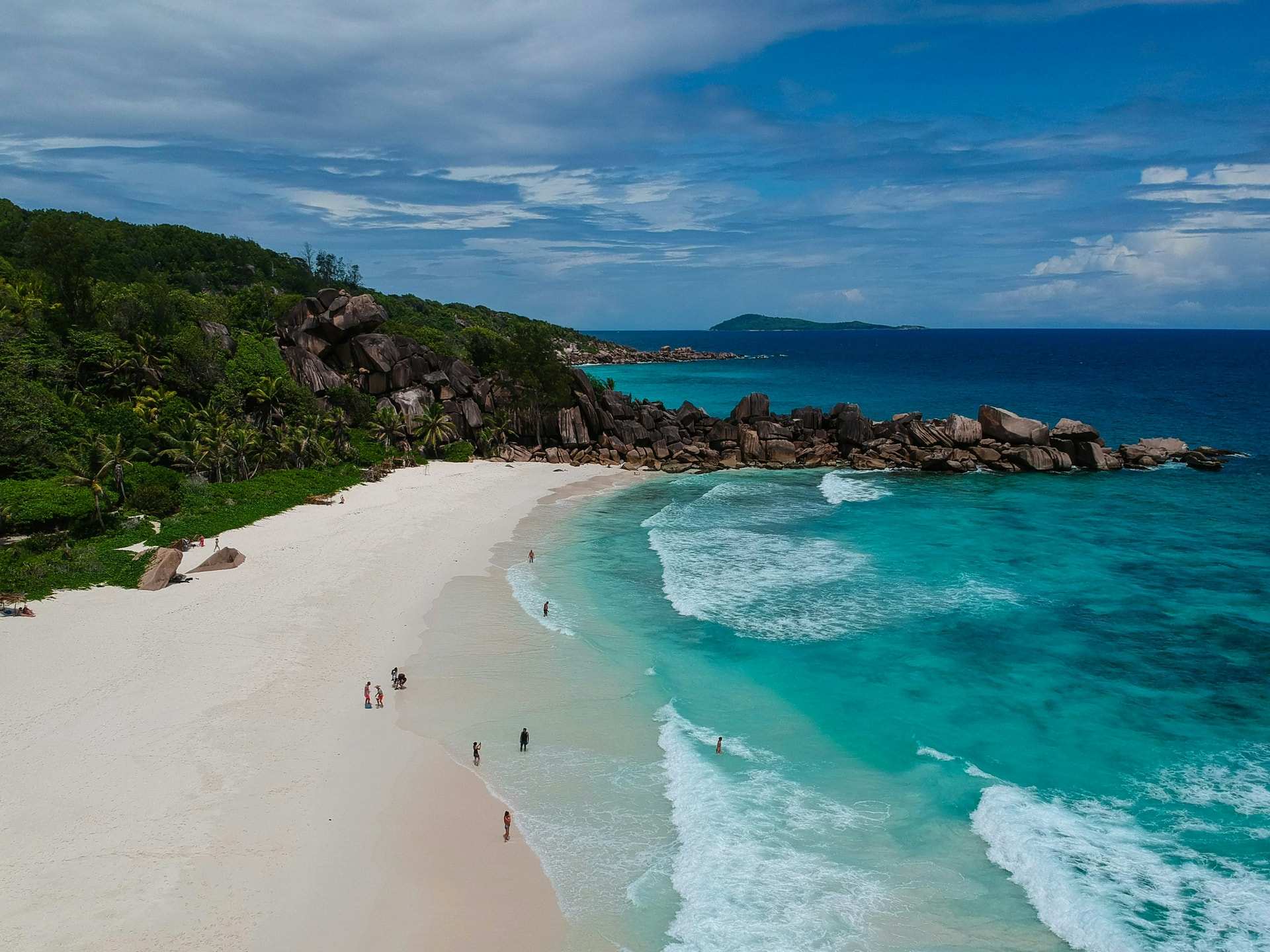 Waves running up a long white beach below green granite hills, a few people walking at the water's edge