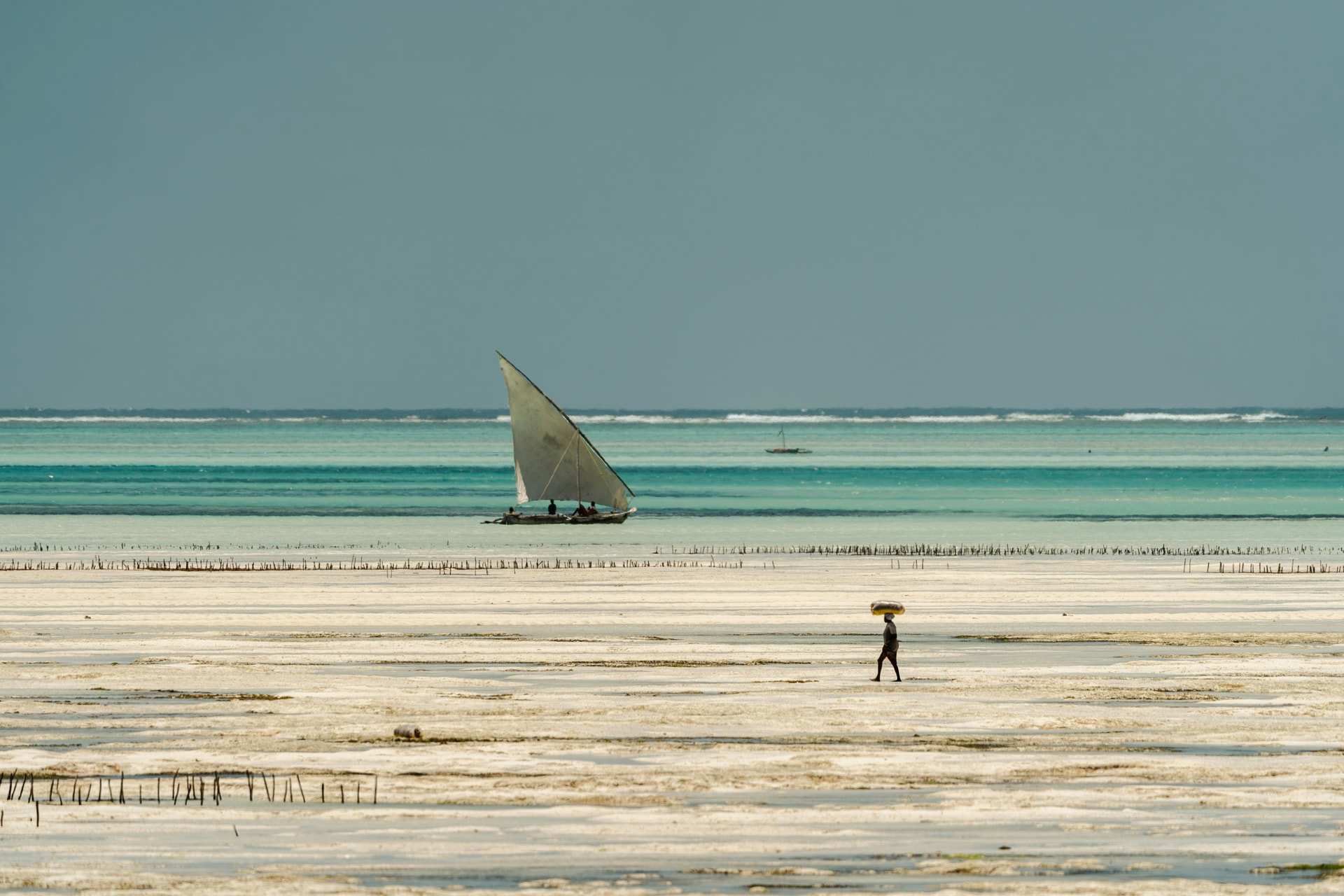 Low tide at Jambiani on Zanzibar's east coast, the sea far out across wet sand, a woman carrying goods along the shore and a dhow under sail beyond the reef