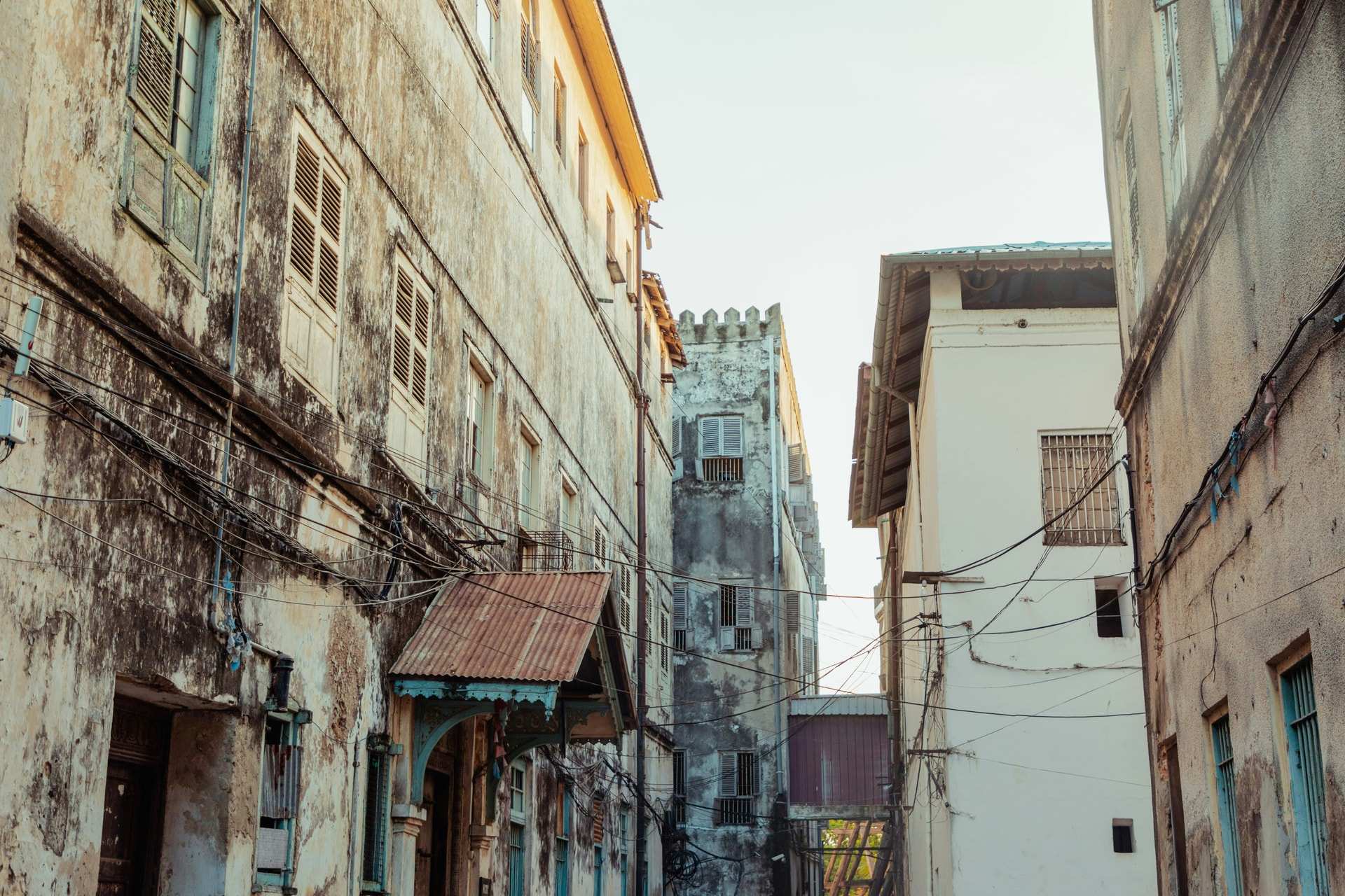 A narrow Stone Town street between weathered coral-stone buildings with shuttered windows, an old tower at the far end