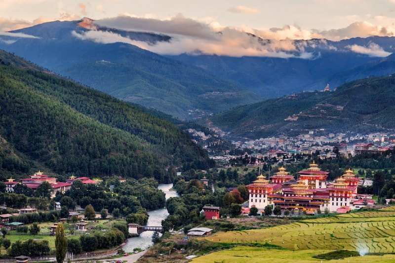 A white-walled dzong with gold-trimmed roofs beside the river in the Thimphu valley, the town and forested mountains behind