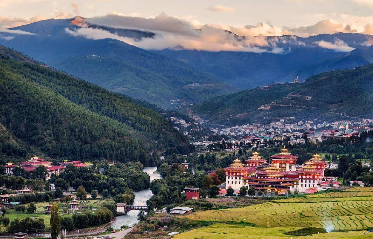A white-walled dzong with gold-trimmed roofs beside the river in the Thimphu valley, the town and forested mountains behind