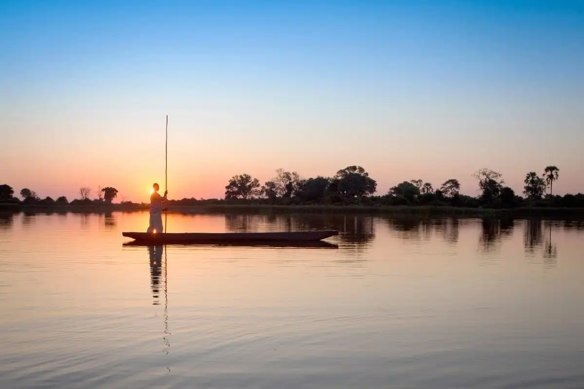 A poler guiding a dugout mokoro across still water at sunset