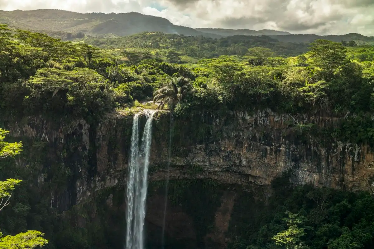 A twin waterfall dropping into a forested gorge below green hills