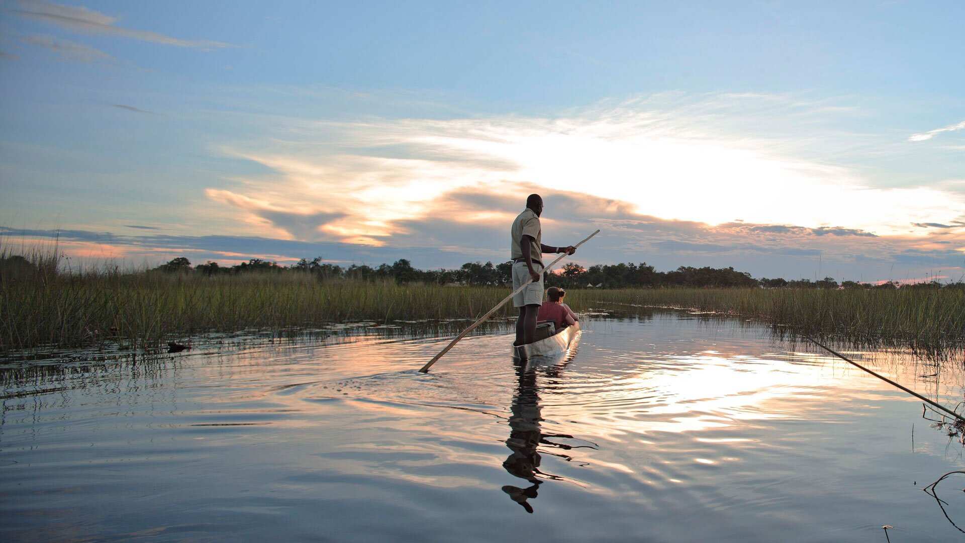 A guide poles a mokoro along a still reed channel in the Okavango Delta at dusk, the sky mirrored in the water