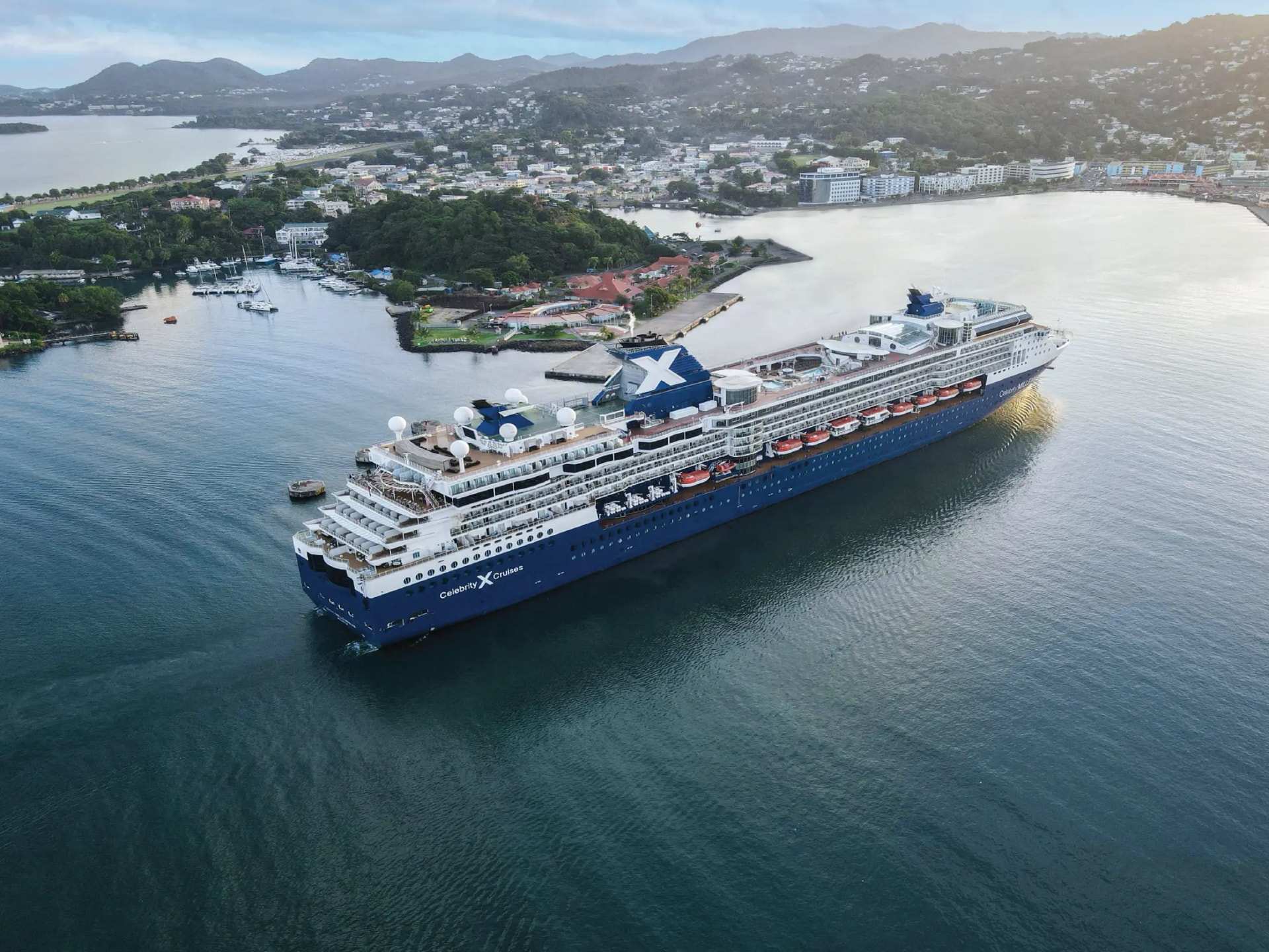 A Celebrity ship at anchor in a bay below a green hillside town