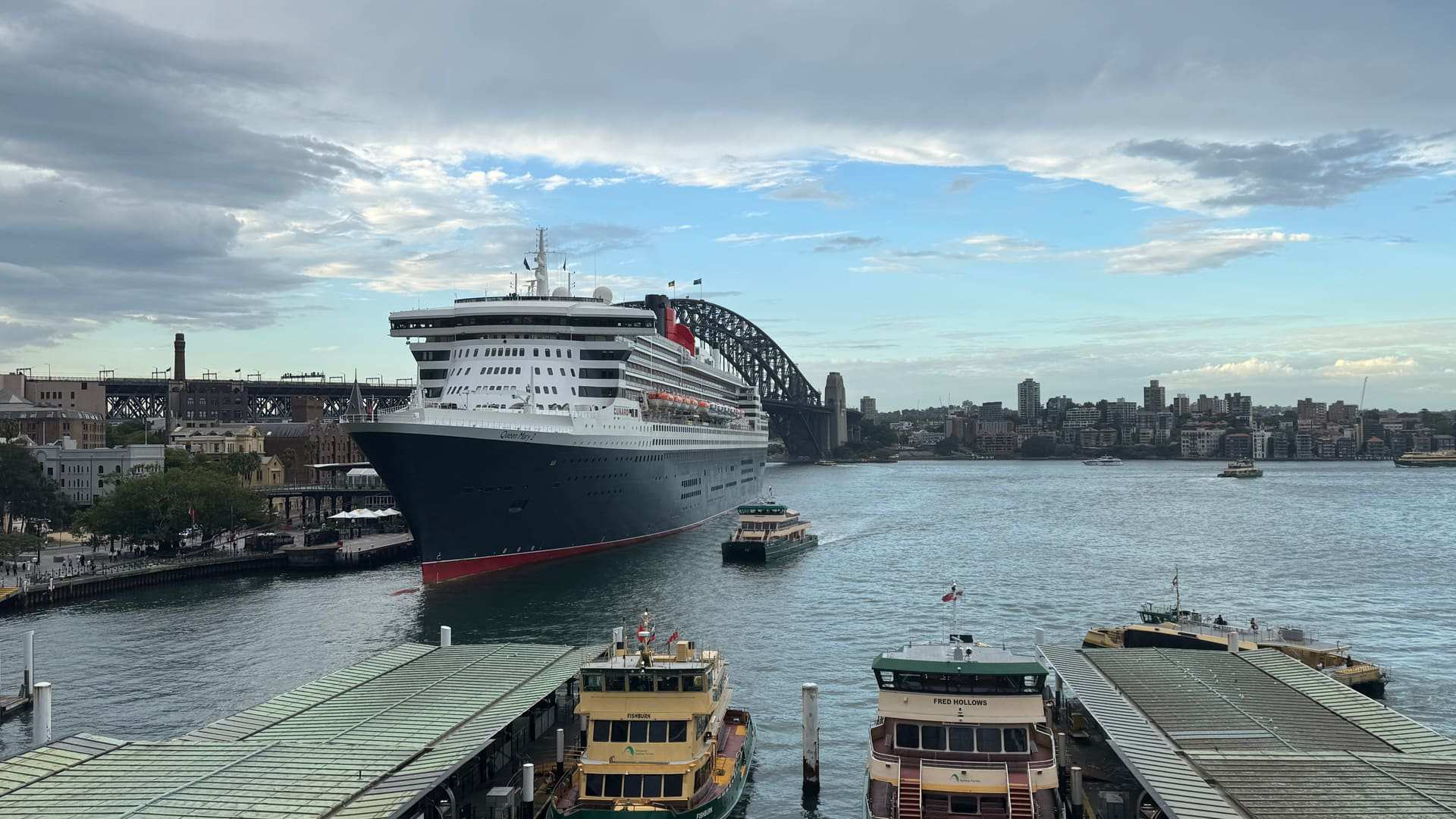 Queen Mary 2 berthed in Sydney Harbour, the Harbour Bridge behind her and ferries at the wharves in front