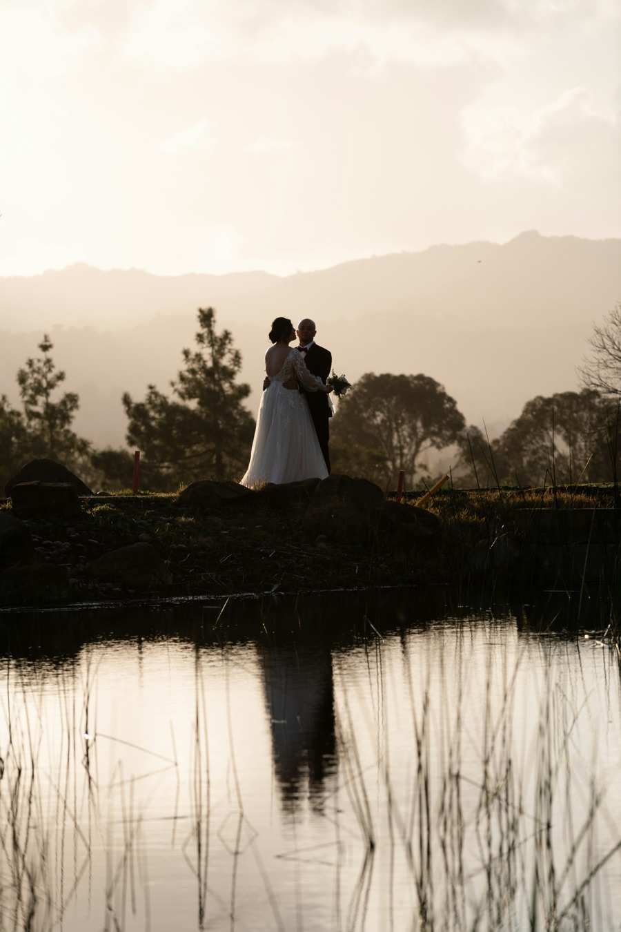 A couple in wedding clothes on a rock above still water, hills behind them at dusk