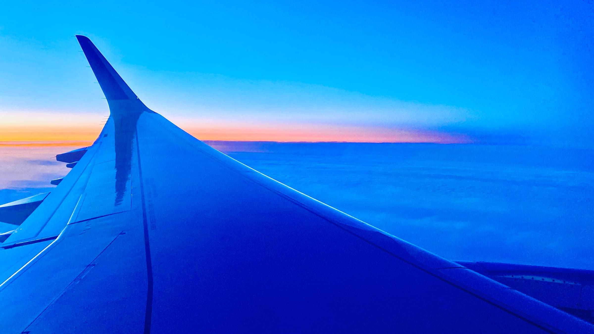 An airliner's wing at dusk from a passenger window, level above a blue cloud deck, the sunset still orange along the horizon