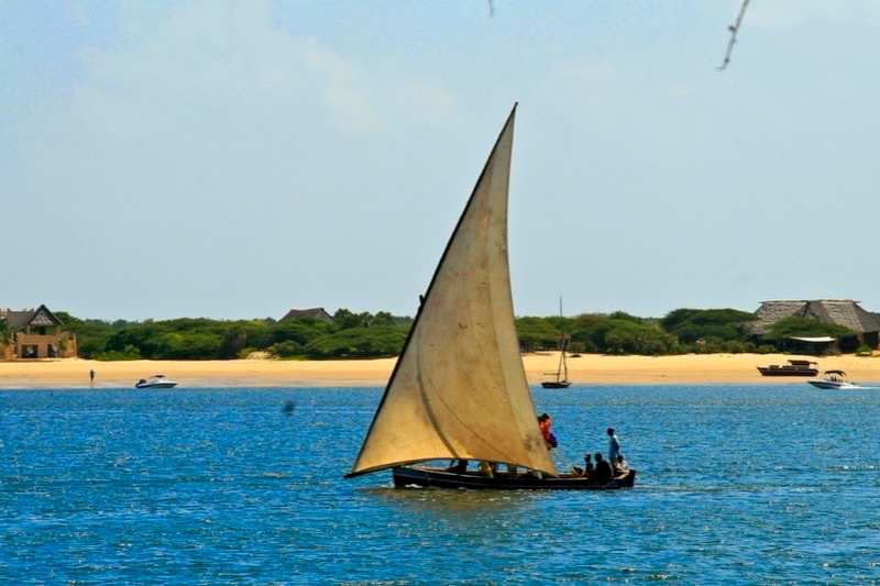A traditional dhow under full sail off Lamu, a white sandbar and thatched roofs along the shore behind it