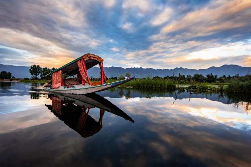 A canopied shikara on Dal Lake in Srinagar, boat and cloudy sky mirrored in still water, mountains beyond