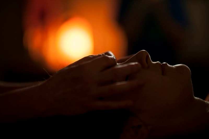 Hands working across a person's face during a massage, a warm lamp glowing in the dark behind