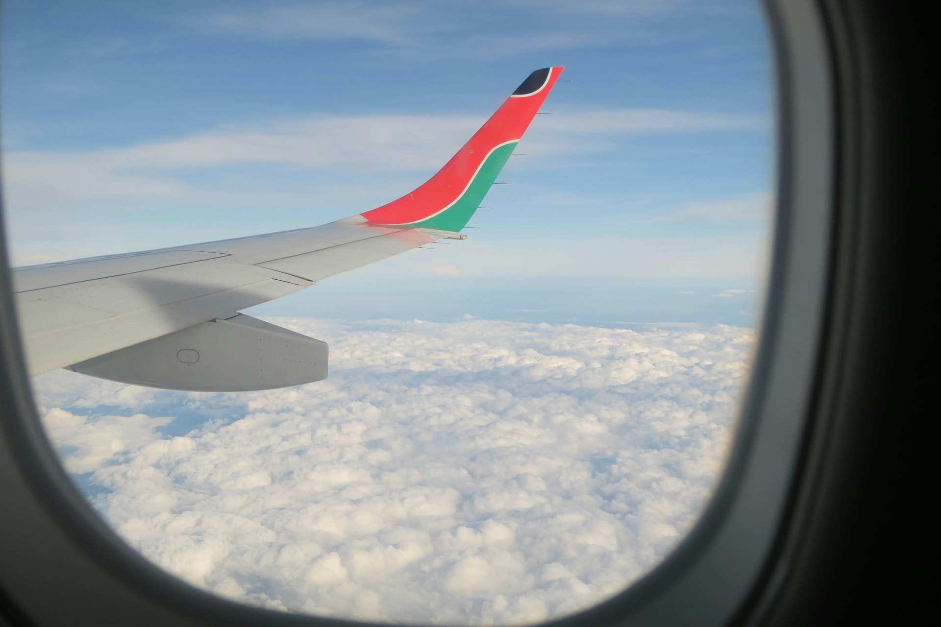 A Kenya Airways wingtip in red, green and black seen through a cabin window above a layer of white cloud