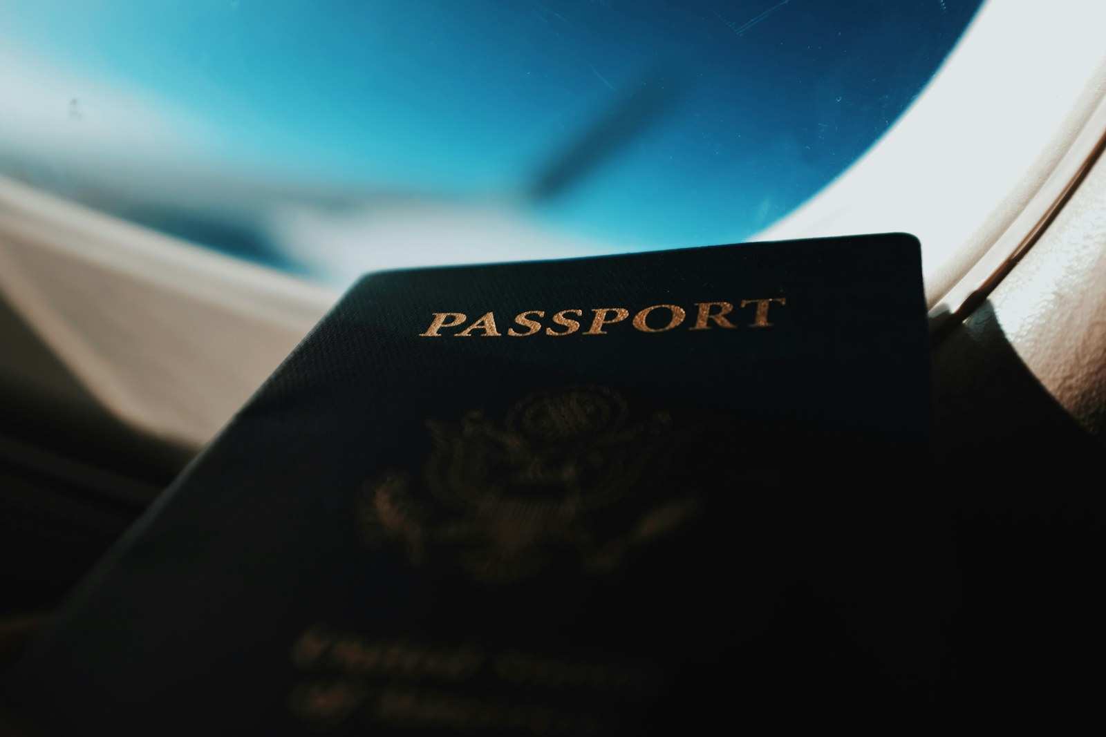 A dark passport held up against an aircraft cabin window, the wing and a bright blue sky behind it