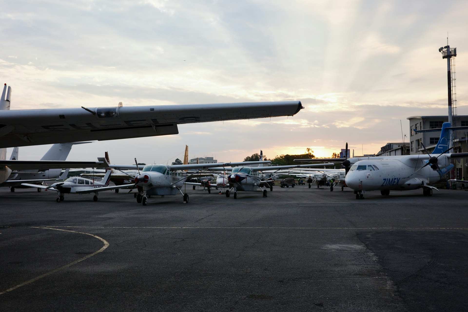 Light aircraft and a small turboprop parked on the apron at Wilson Airport, Nairobi, with the sun low behind a hangar