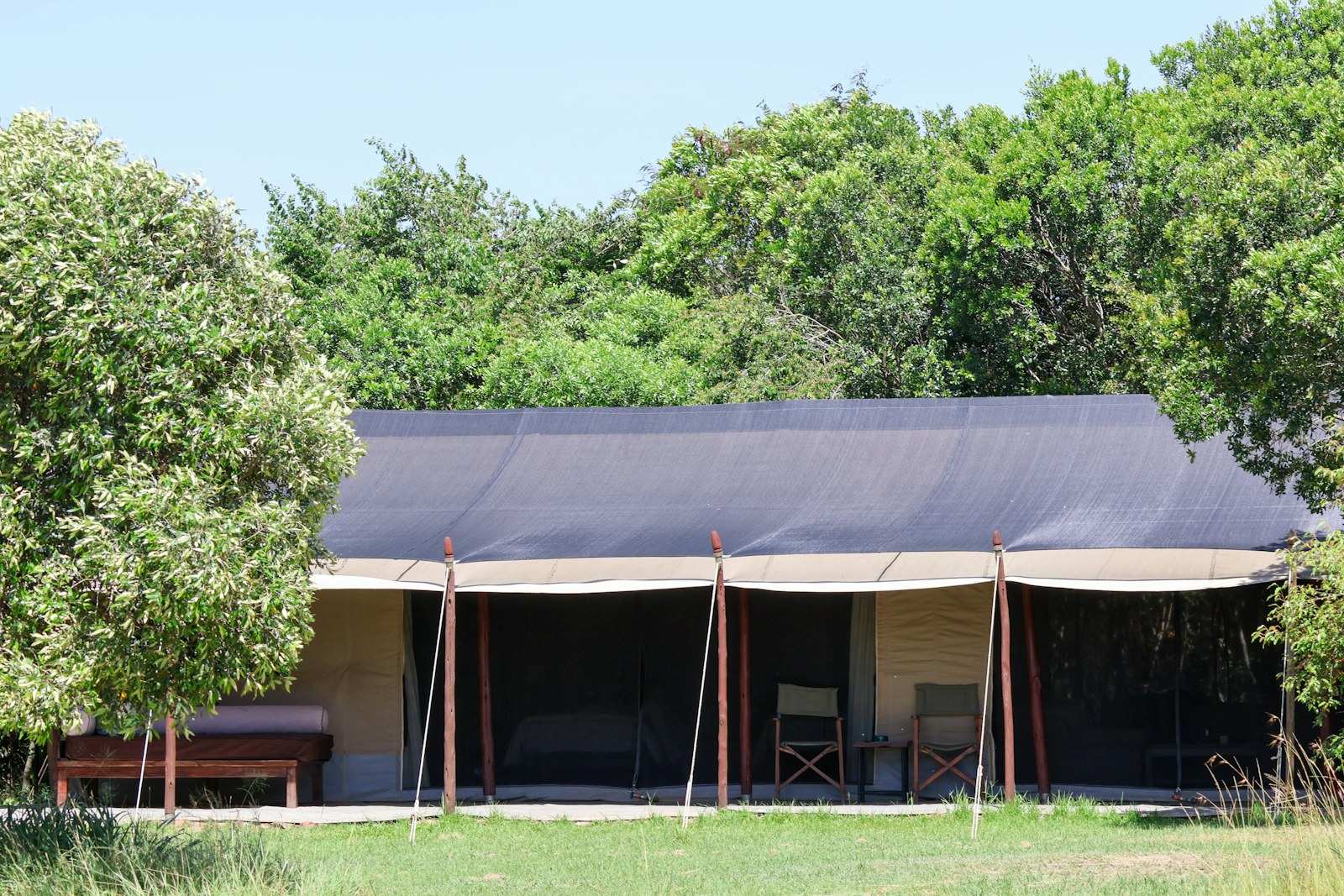 A safari tent under a dark canvas fly, its mesh front open on a bed, two folding chairs and a low table, with a day bed on the grass outside