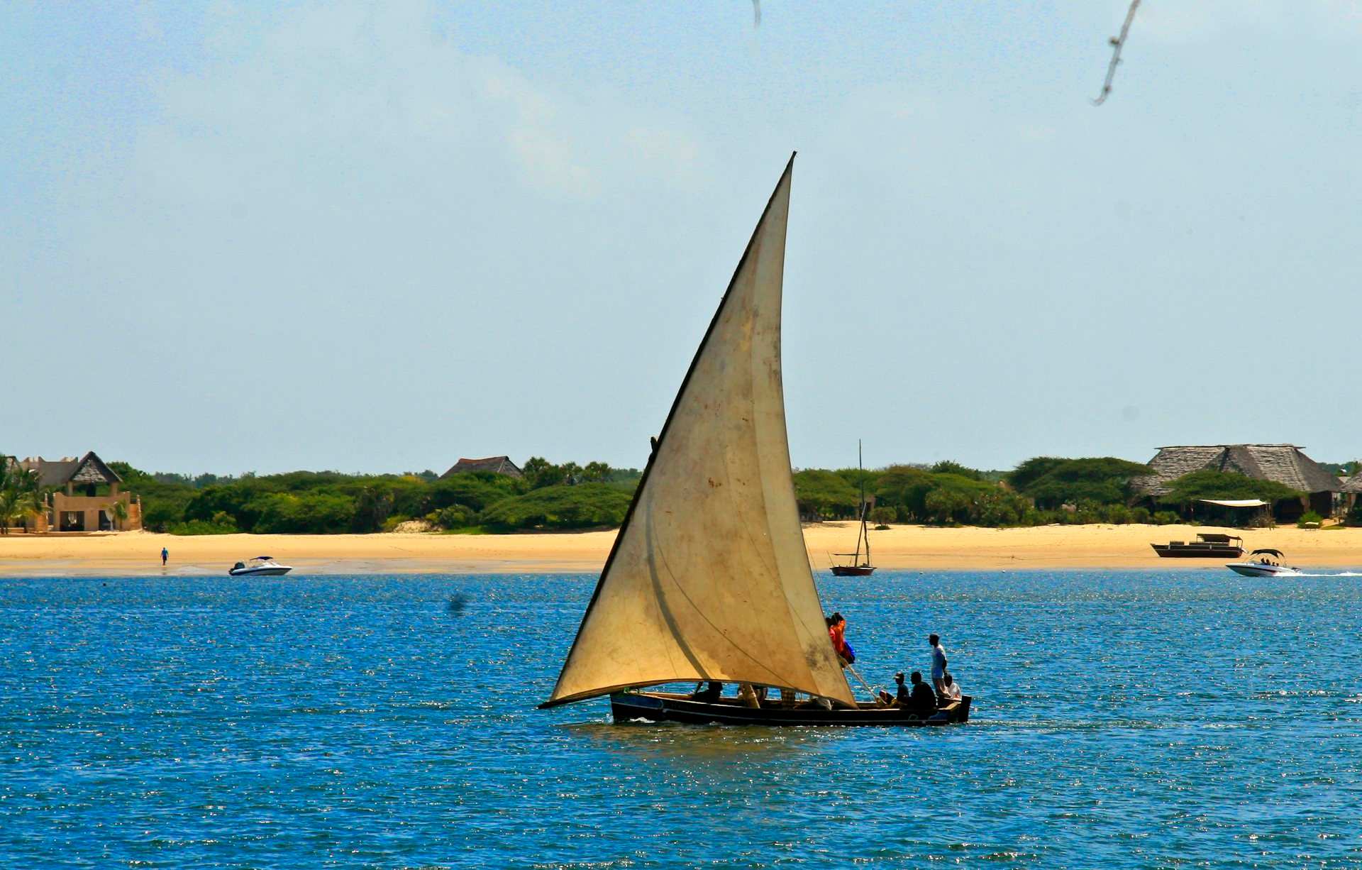 A traditional dhow under full sail off Lamu, a white sandbar and thatched roofs along the shore behind it