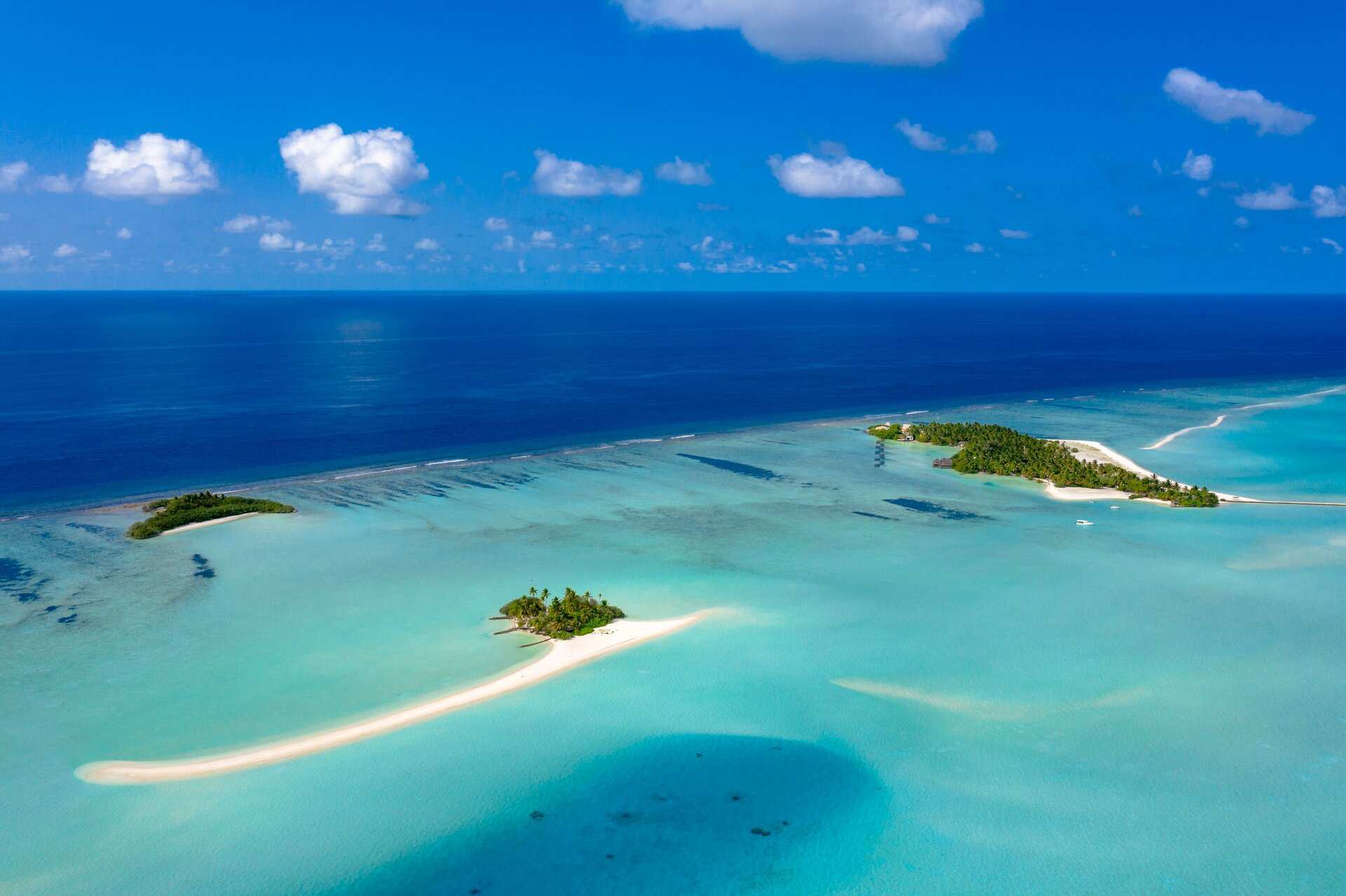 An aerial view of a Maldivian atoll, with small palm-covered islands and a curving sand spit in a turquoise lagoon, and the reef edge dropping to deep blue ocean