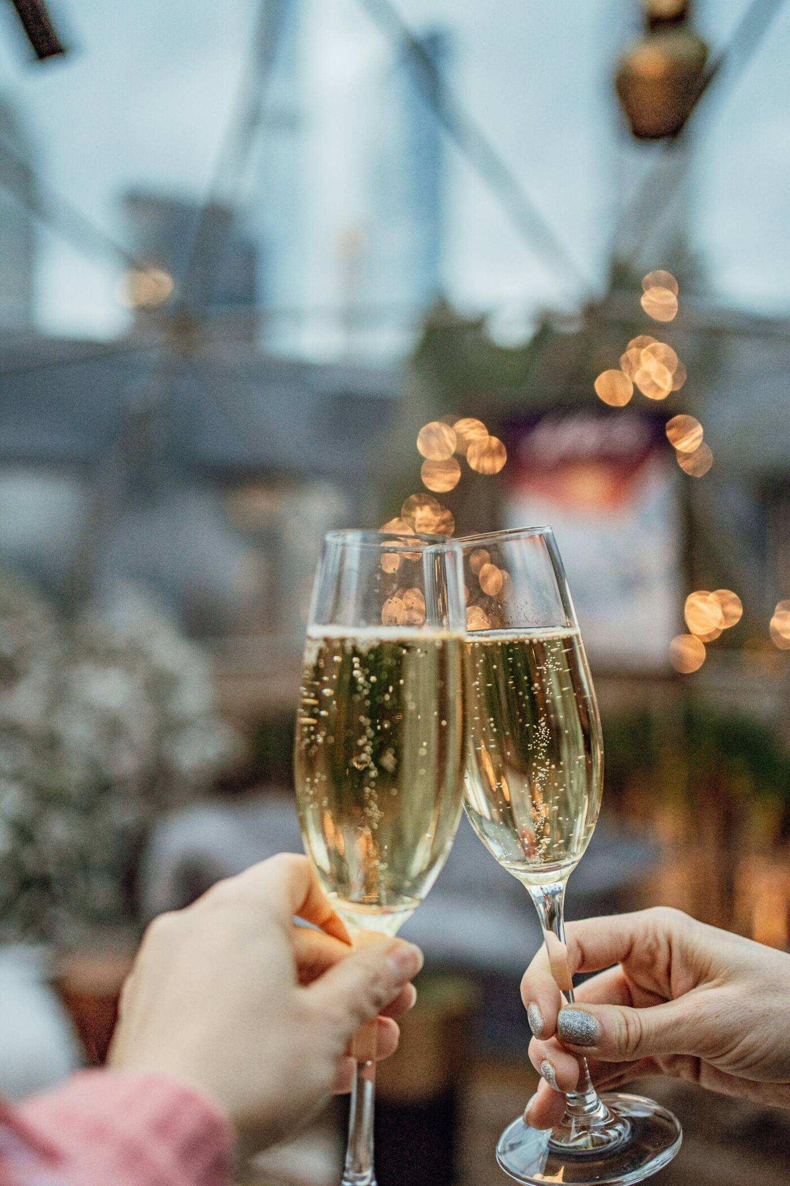 Two people touching champagne glasses together at dusk, strung lights glowing behind them