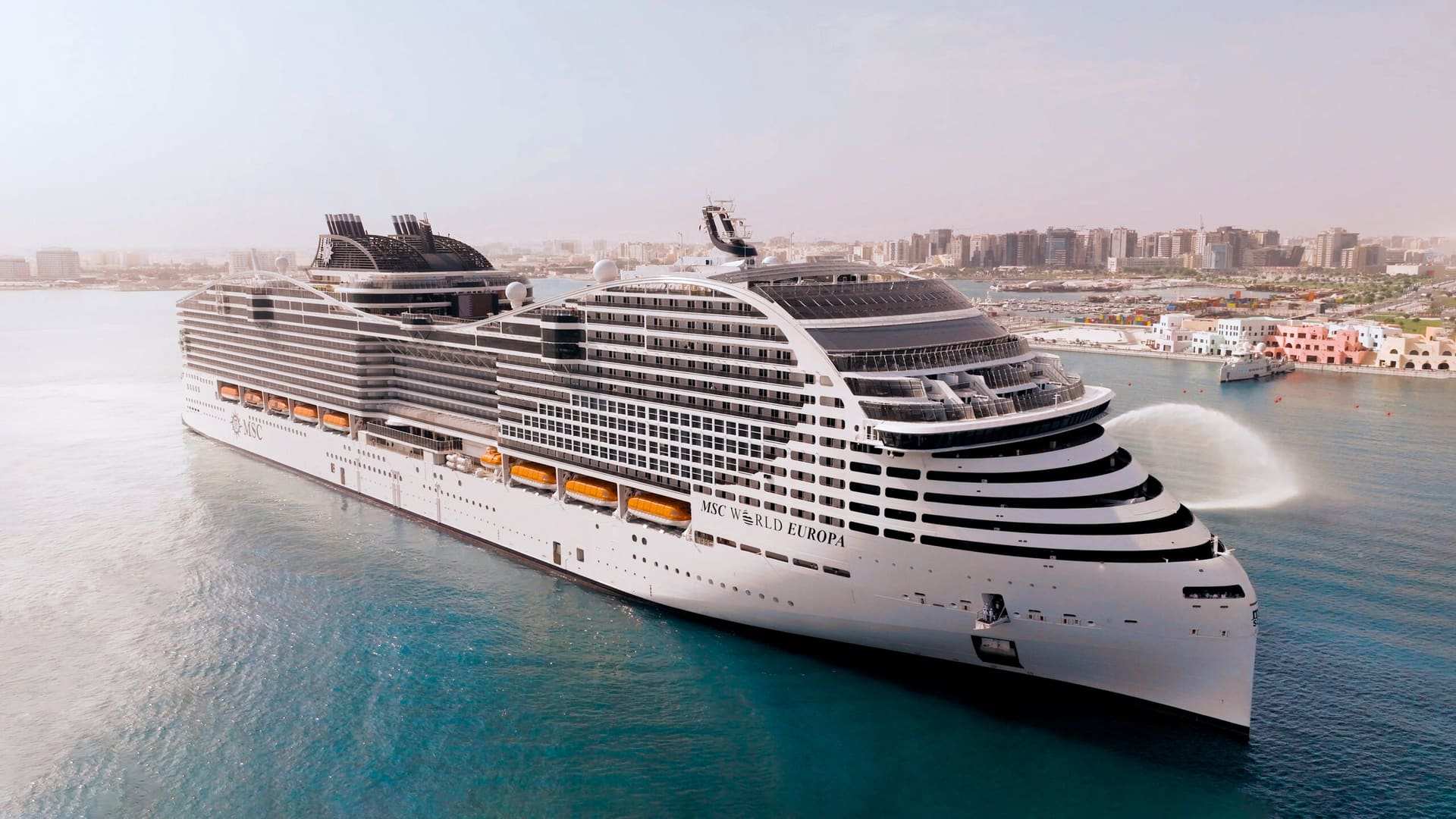 MSC World Europa under way off a city waterfront, her name on the hull and a plume of spray arcing beside her bow