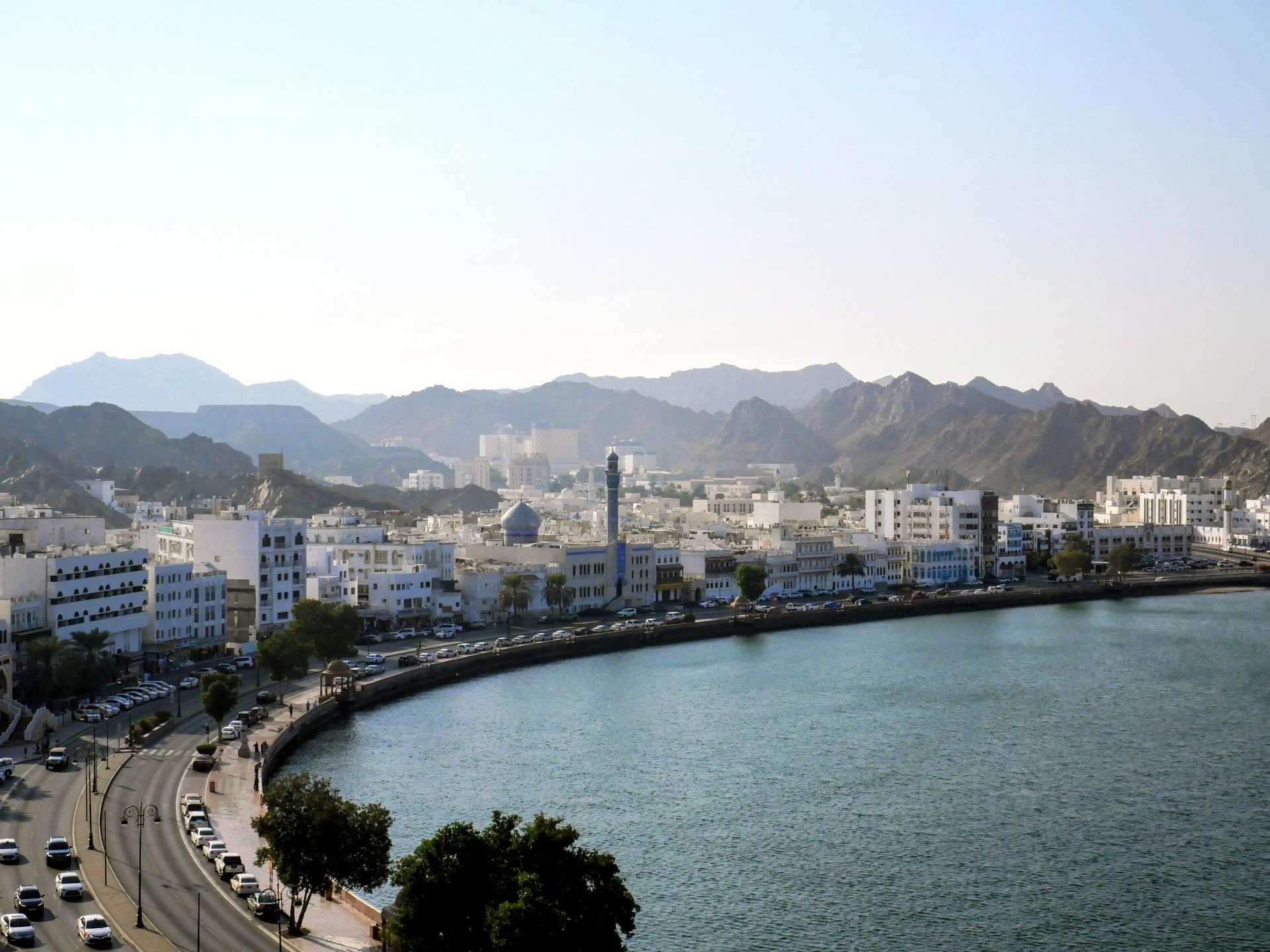The Muttrah Corniche curving around the harbour in Muscat, white buildings along the waterfront and the bare ridges of the Hajar Mountains behind