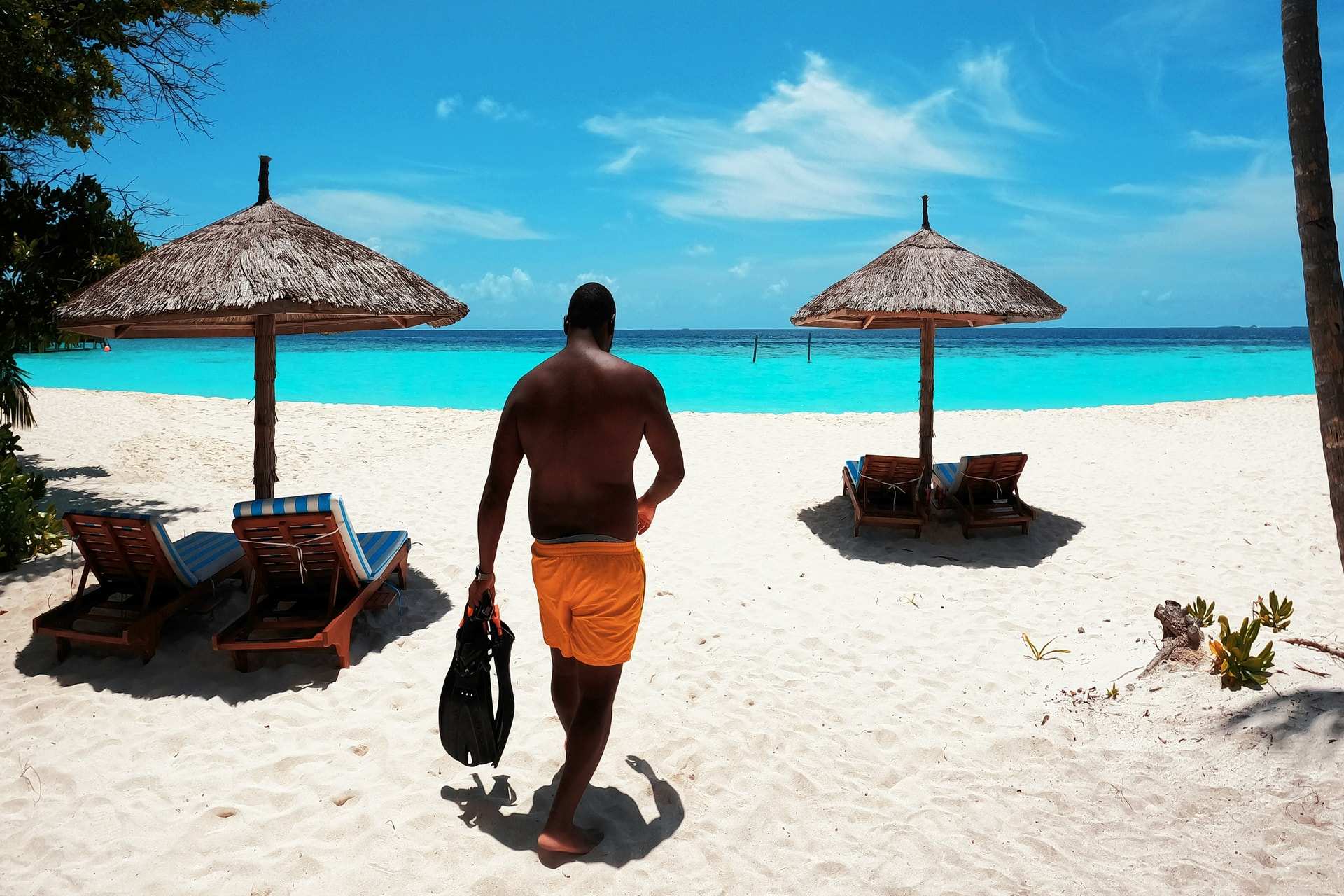 A man in orange swim shorts walks across white sand towards two thatched sunshades and a turquoise lagoon in the Maldives, carrying a pair of fins