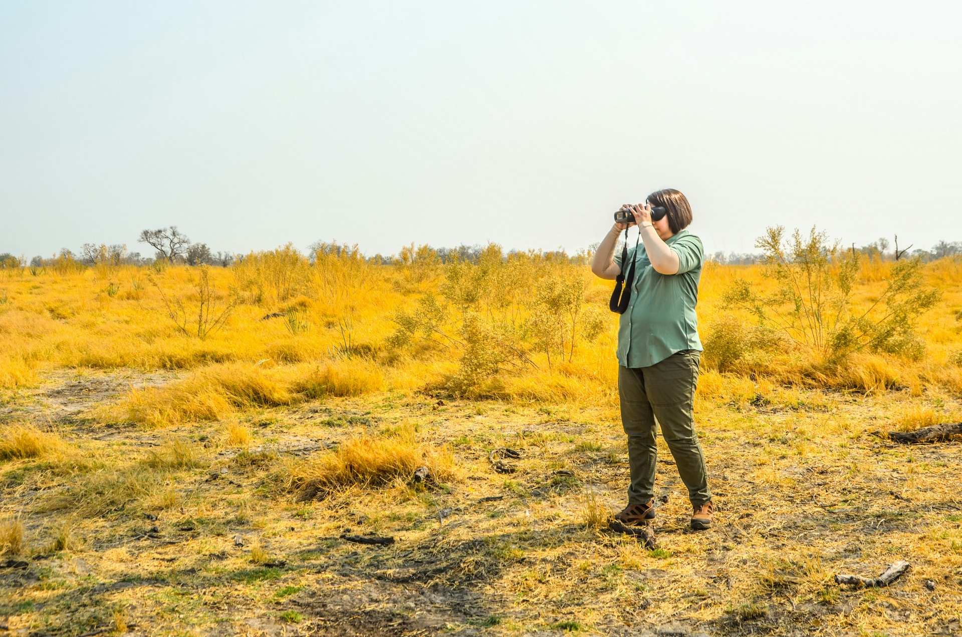 A traveller in a green shirt and khaki trousers raising binoculars to scan open golden grassland under a pale sky
