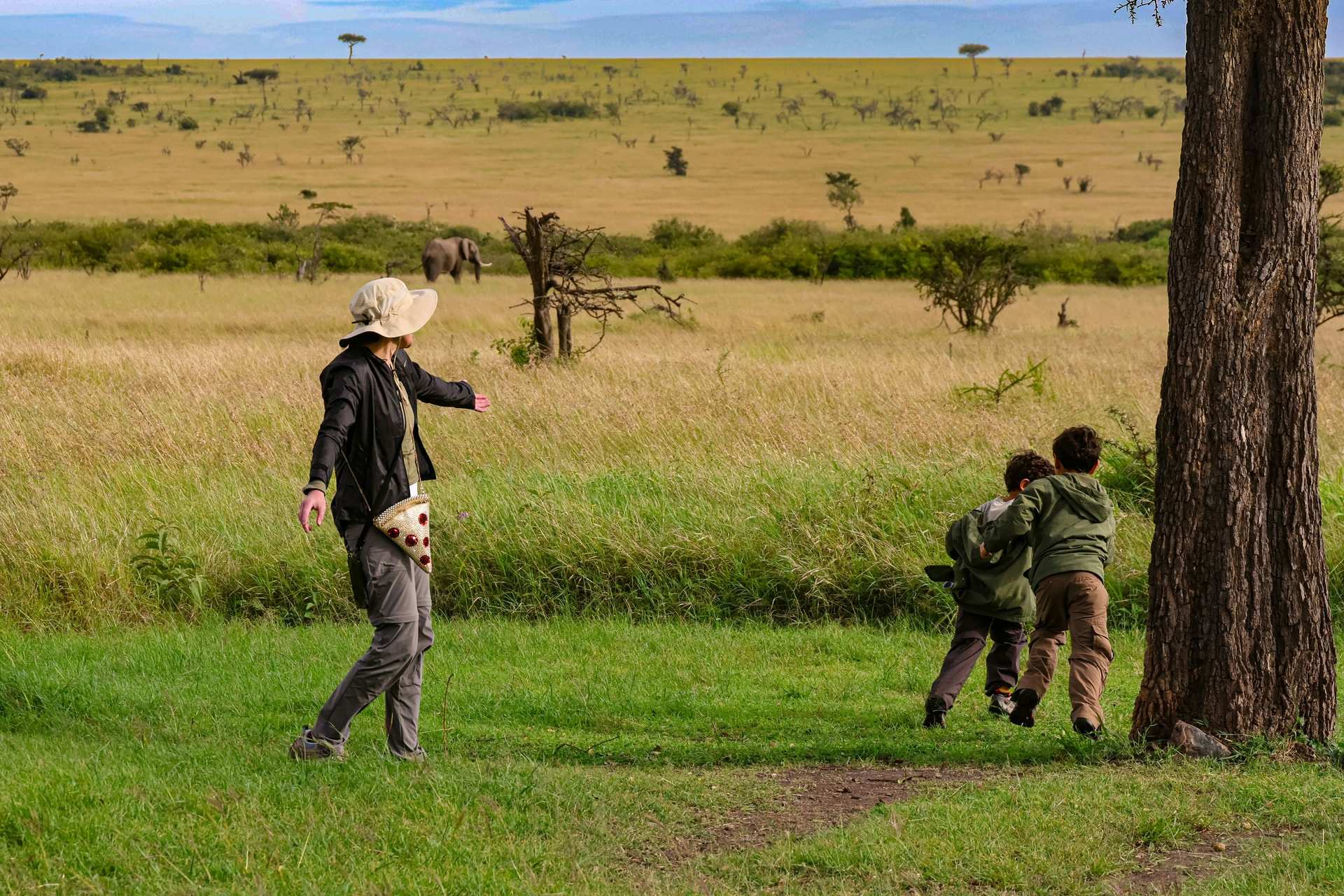 Two small children in green jackets run across short grass beside a tree trunk while an adult in a sun hat follows, with an elephant on the plains behind them in the Naboisho Conservancy