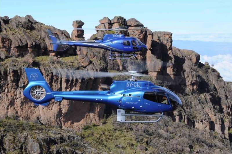 Two blue Tropic Air helicopters flying in formation past red rock pinnacles high above the cloud
