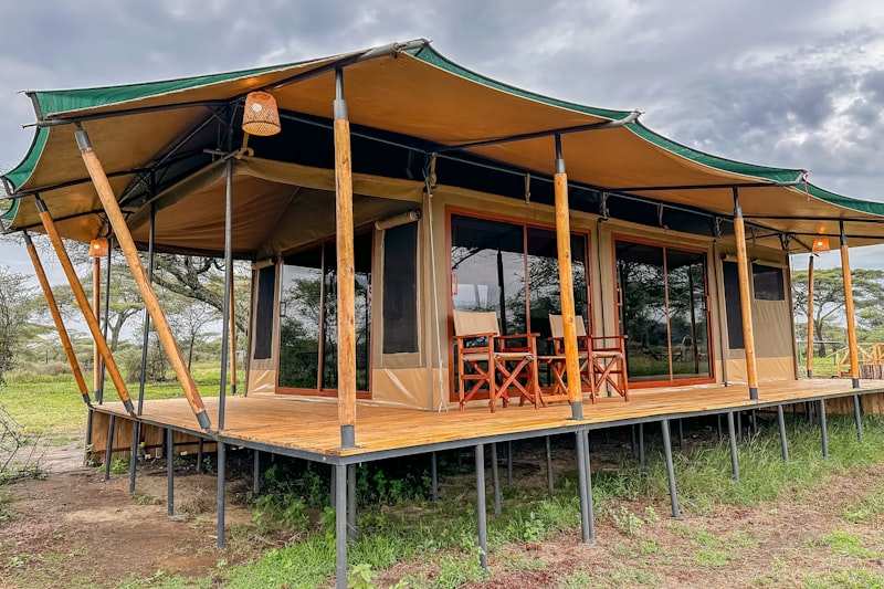 A canvas safari tent on a raised deck, directors' chairs on the veranda and acacia beyond