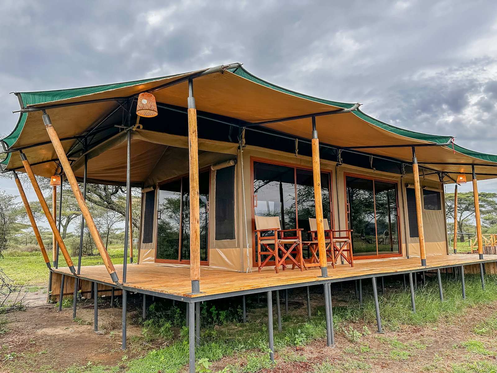 A canvas safari tent on a raised deck, directors' chairs on the veranda and acacia beyond