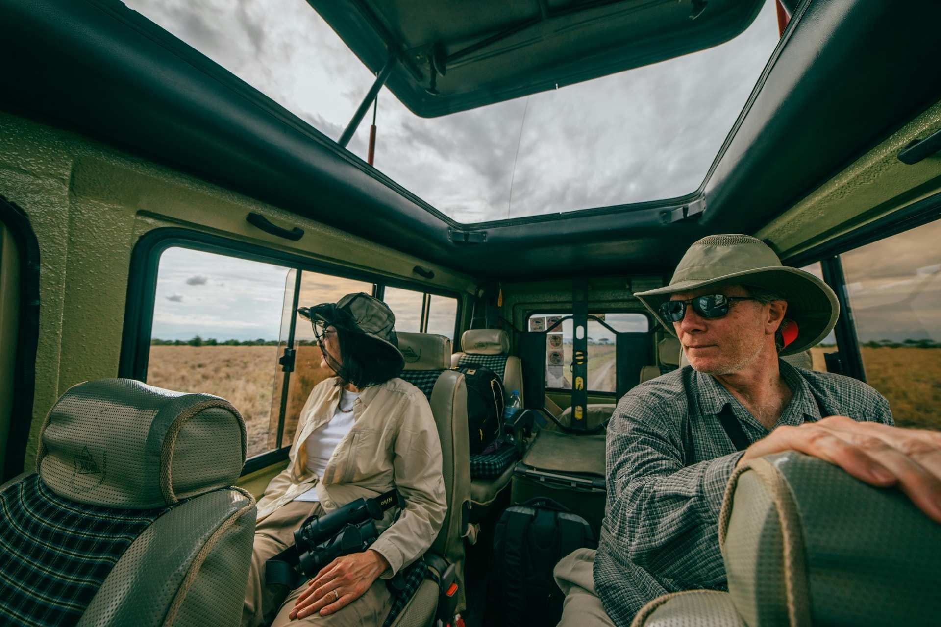 Two guests seated inside a pop-top safari vehicle, the canvas-lined roof raised above them and open grassland visible through the windows