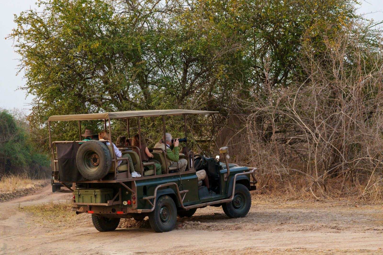 Guests photographing from an open-sided game viewer with a canvas roof, a second vehicle ahead on the track