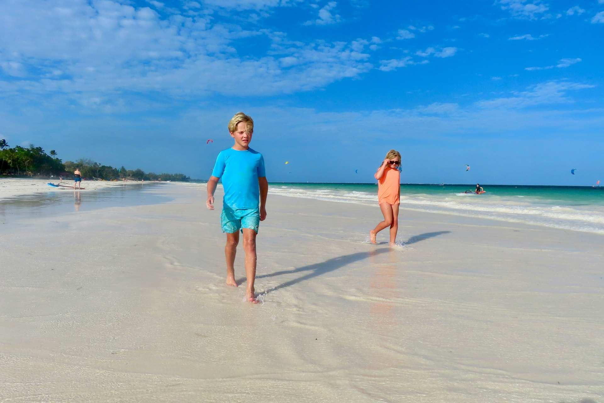 A boy and a girl walk barefoot across wet sand at low tide on Diani Beach, Kenya, under a blue sky