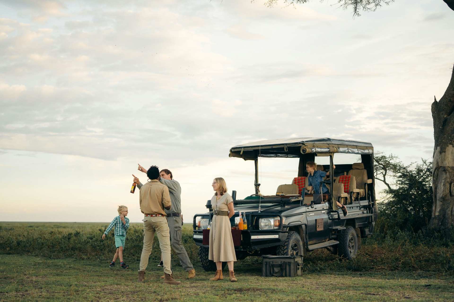 A family beside an open safari vehicle on the grass plains of Tanzania, one adult pointing across the plain, a small boy on the grass and a second child in the back seat