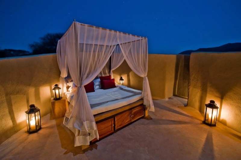 A four-poster bed under mosquito netting on an open terrace, lit by lanterns at dusk