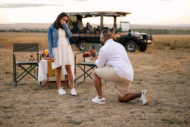 A man on one knee with a ring box, a set table and a safari vehicle behind them on the evening plains
