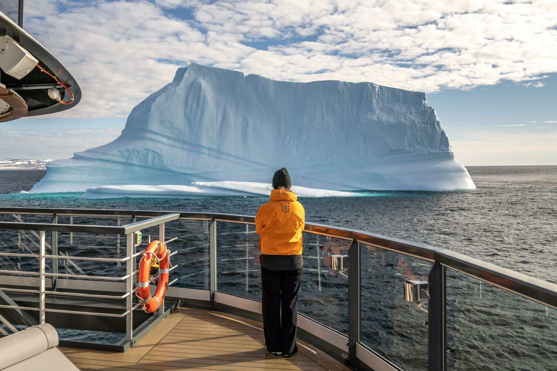 A guest in an orange parka at the deck rail of Seabourn Venture, watching a flat-topped iceberg on a polar sea