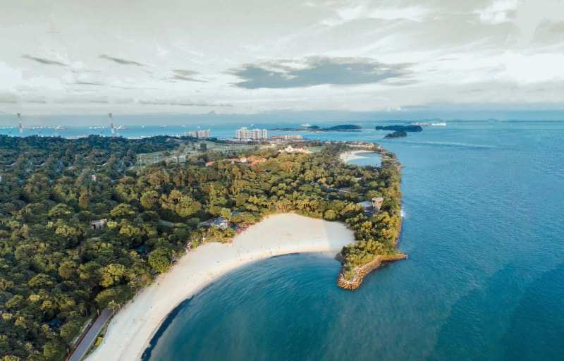 Sentosa Island from the air, a curve of white beach below dense trees and ships out at sea
