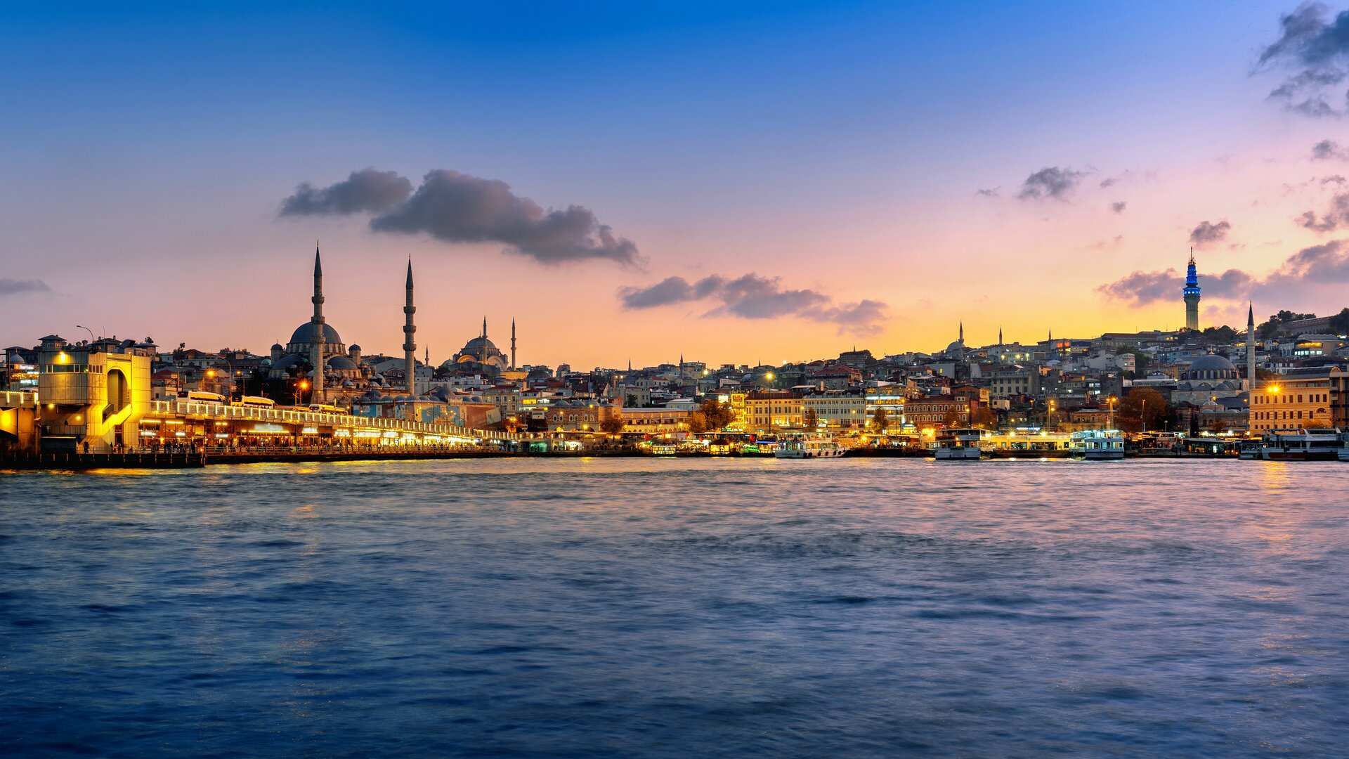 Istanbul's old city at twilight seen across the water, mosque domes and minarets on the skyline above the lit deck of the Galata Bridge