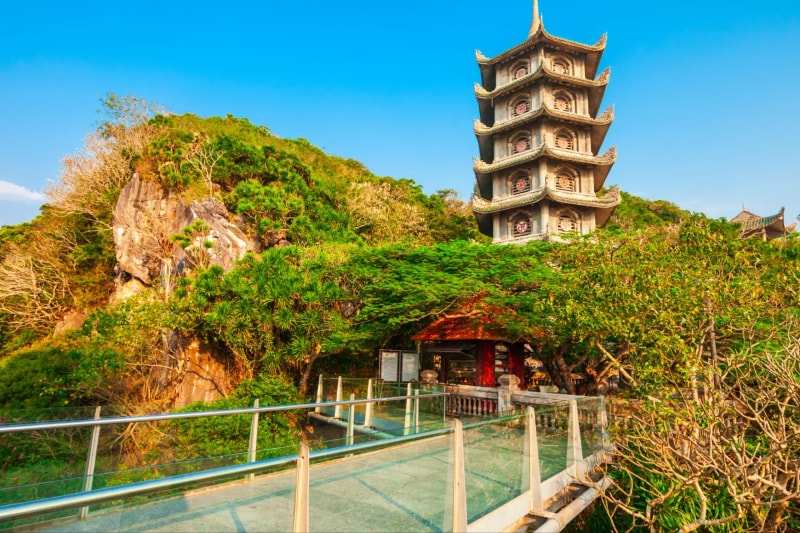 A tiered pagoda tower on a rocky, wooded hilltop, a glass-sided walkway and a small red shrine below