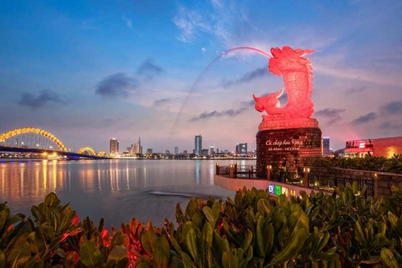 The carp-and-dragon statue on the Han River bank in Da Nang, lit red and spouting water at dusk, the Dragon Bridge glowing gold behind