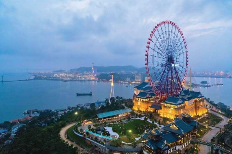 The Sun Wheel lit up at dusk above tiered-roof halls in Ha Long, the bay and the town's lights behind